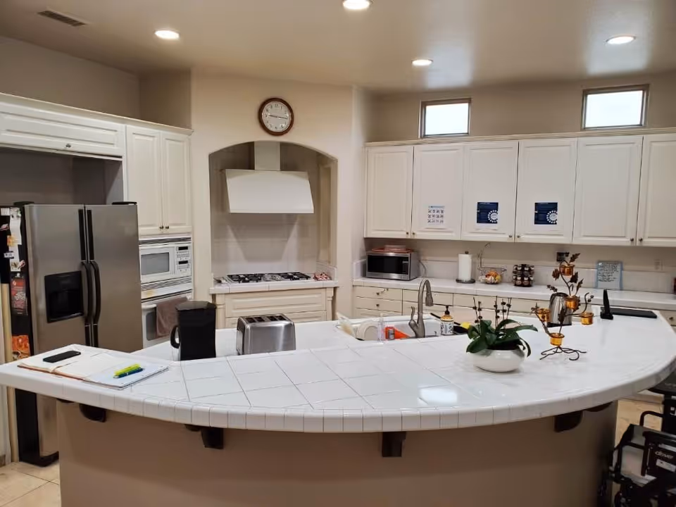 A clean and well-lit kitchen with white cabinets and tiled countertops. The kitchen features a stainless steel refrigerator, a microwave, a gas stove with a white range hood, a toaster, a coffee maker, and a sink. There is a clock on the wall above the stove, some papers and pens on the counter, and a decorative plant and candle holder on the curved countertop.