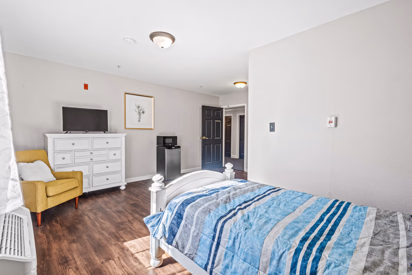 A bright bedroom with a white wooden bed covered in a blue and gray striped comforter. There is a yellow armchair with a white pillow next to a white dresser with a flat-screen TV on top. A small black microwave and mini fridge are positioned against the wall near an open black door leading to a hallway. The room has wood flooring and light-colored walls with two ceiling light fixtures.