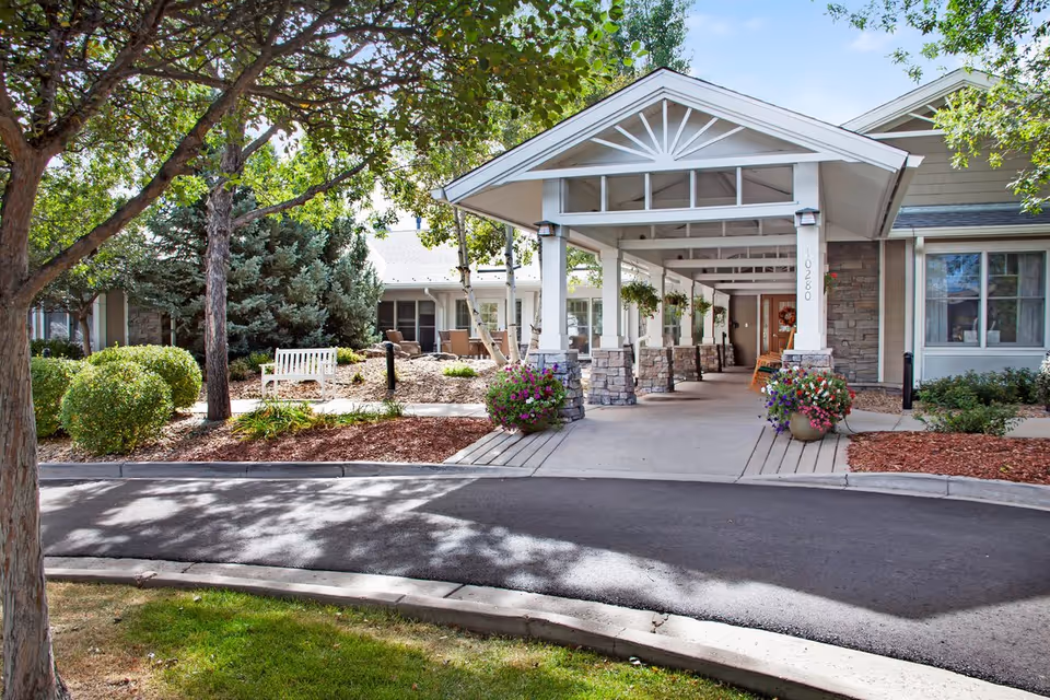 Entrance of a senior living facility with a covered driveway supported by stone pillars, surrounded by landscaped greenery including trees, bushes, and flower pots. A white bench is visible on the left side near the garden area.