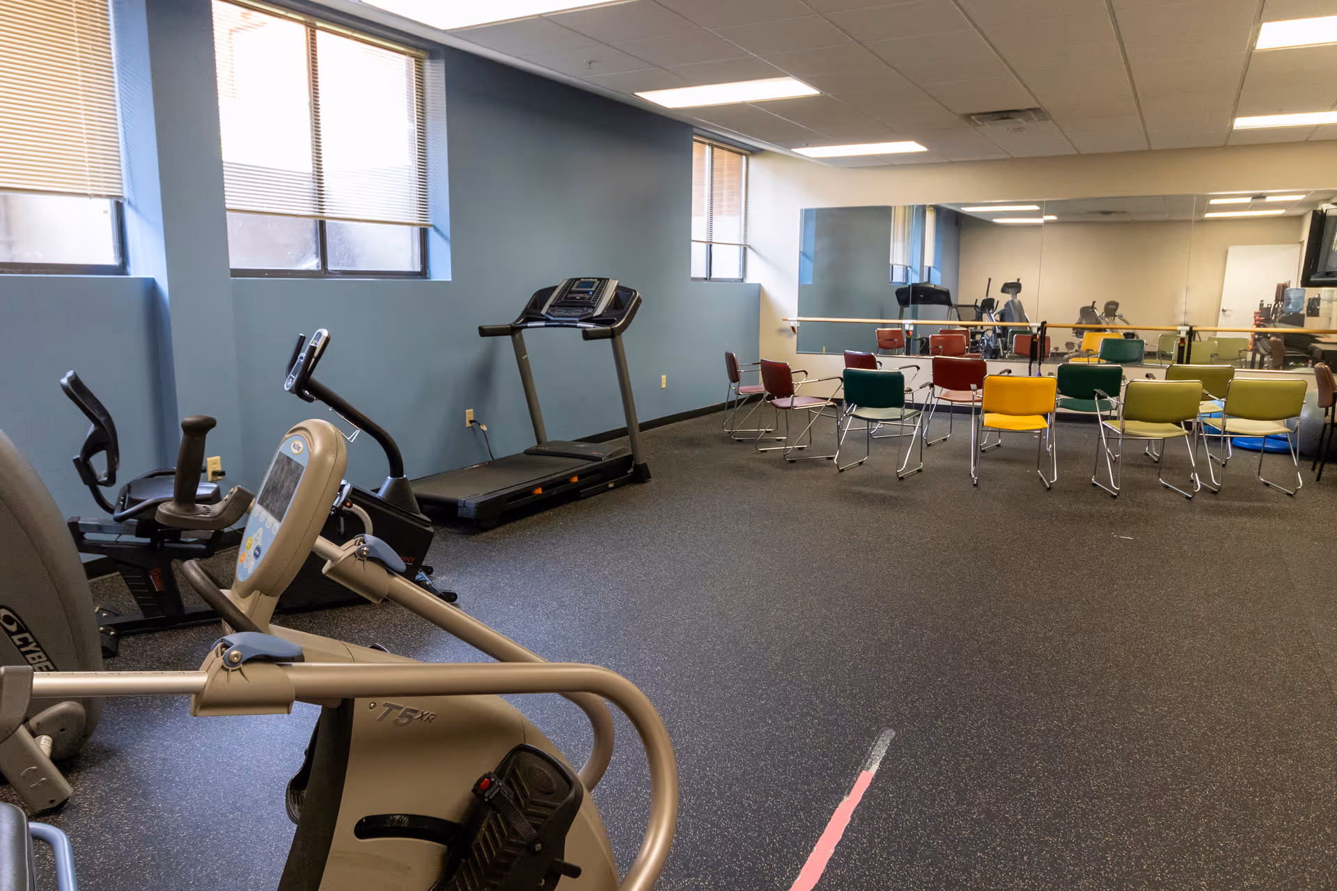 Exercise room with treadmills and stationary bikes and rows of colorful chairs facing a mirrored wall.