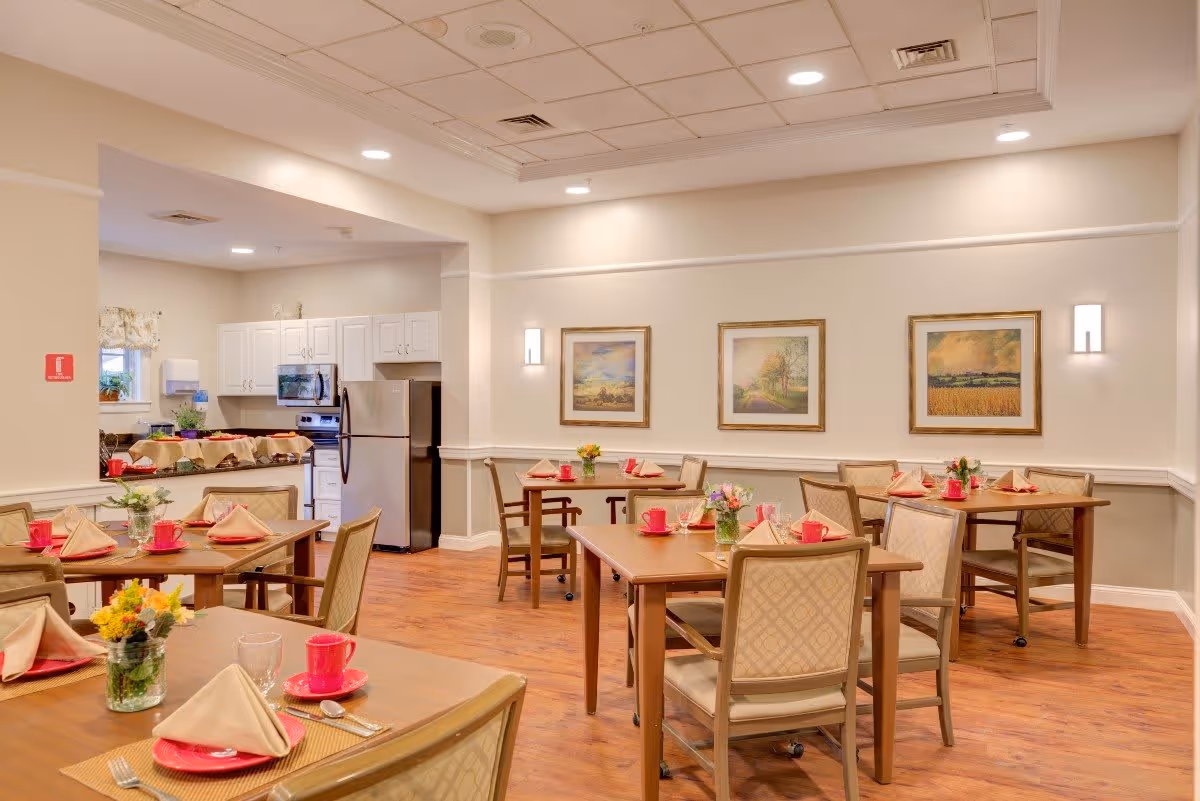 A dining room in a senior living facility with several wooden tables set with red cups, beige napkins, and small flower arrangements. The room has wooden flooring, cream-colored walls with three framed landscape paintings, and recessed ceiling lights. In the background, there is a kitchen area with white cabinets, a stainless steel refrigerator, and a microwave.