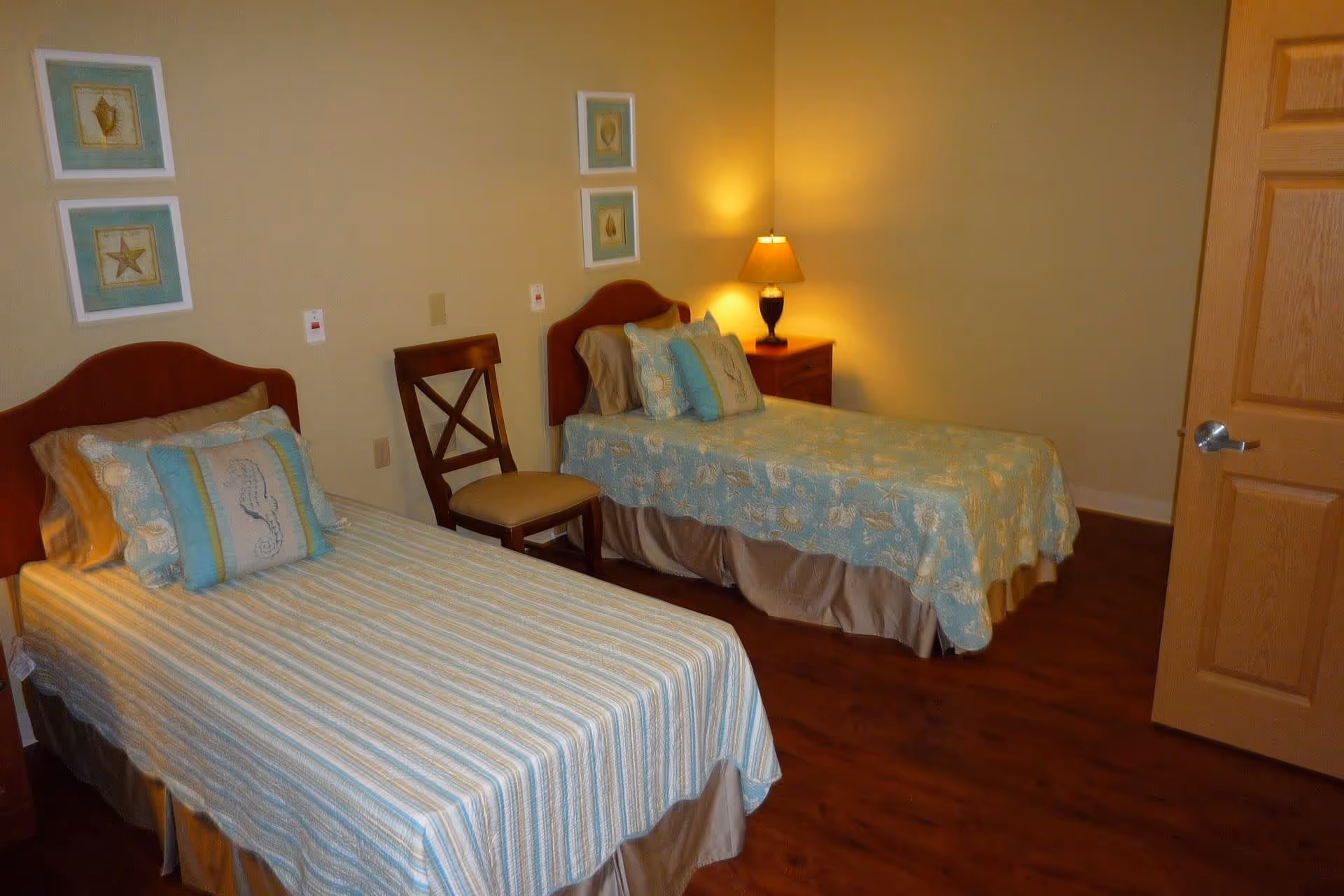 A bedroom with two twin beds, each with a wooden headboard and light blue patterned bedding. There is a wooden chair between the beds and a wooden nightstand with a lamp on it. The walls are beige with two framed pictures above each bed. The floor is wooden, and a wooden door is partially open on the right side of the image.