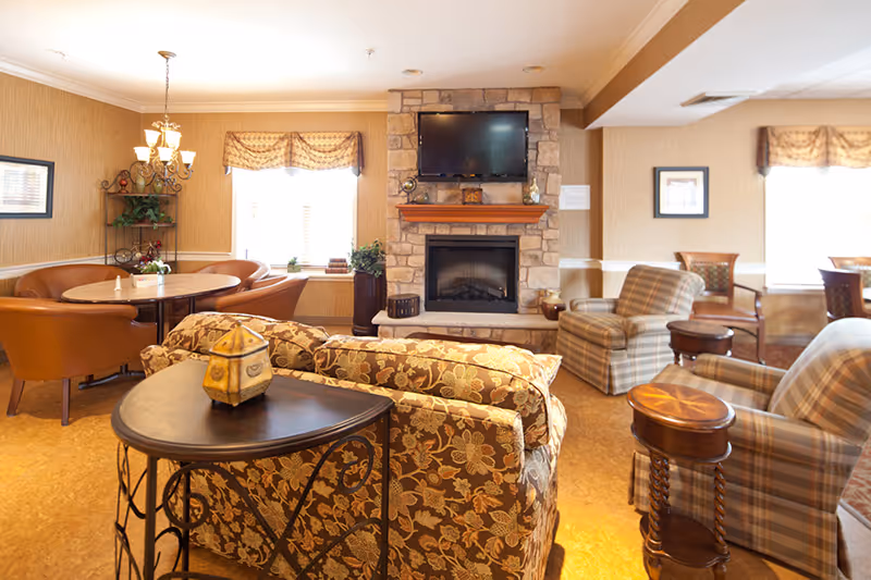 A cozy living room area in a senior living facility featuring a stone fireplace with a mounted flat-screen TV above it. The room includes patterned upholstered sofas and armchairs, wooden side tables, a round dining table with leather chairs, and windows with decorative valances letting in natural light.