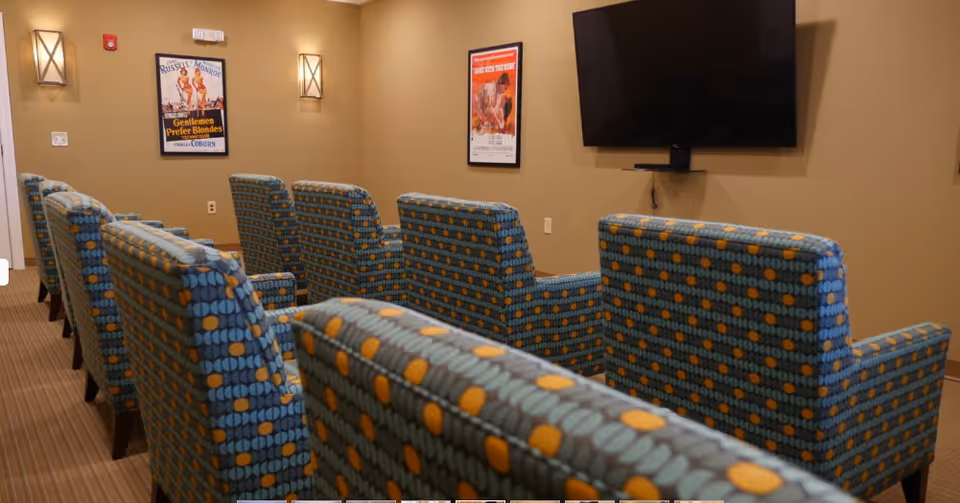 Rows of patterned armchairs facing a wall-mounted TV and framed movie posters in a small media room.