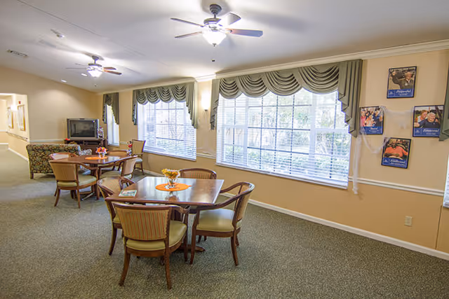 A well-lit common area in an assisted living facility with two wooden tables surrounded by chairs. Large windows with green valances let in natural light. A TV and couch are visible in the background along a beige wall. The carpet is green, and there are framed pictures on the wall near the windows.