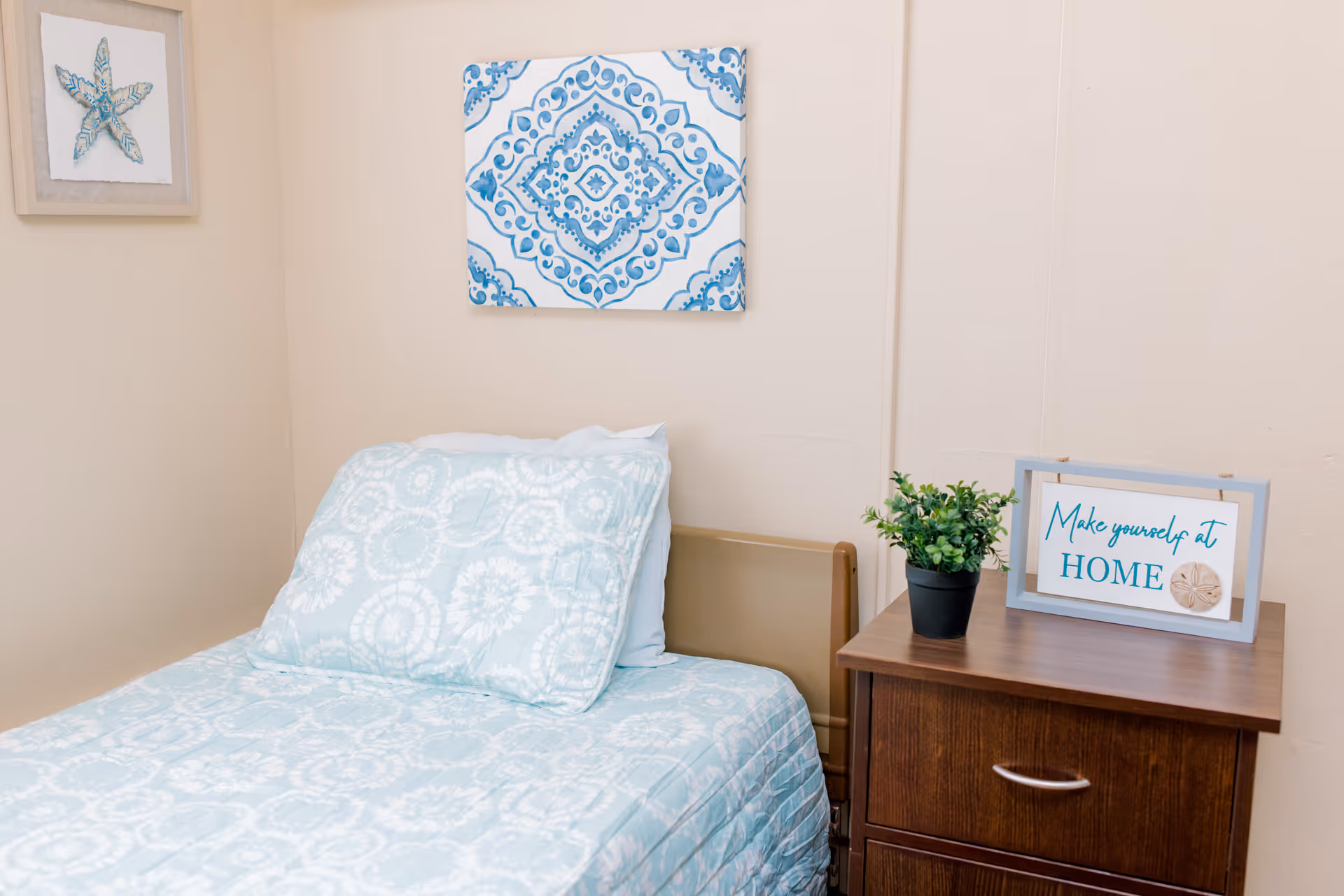 A single bed with light blue patterned bedding beside a wooden nightstand topped with a potted plant and a framed sign reading 'Make yourself at HOME'.