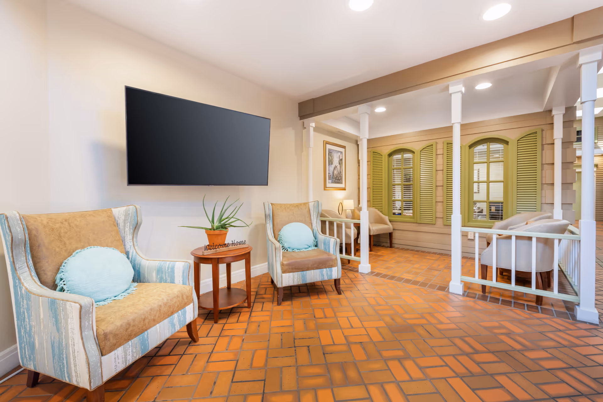 Bright lounge area with armchairs, a wall-mounted TV, and a cozy seating nook with decorative shutters.