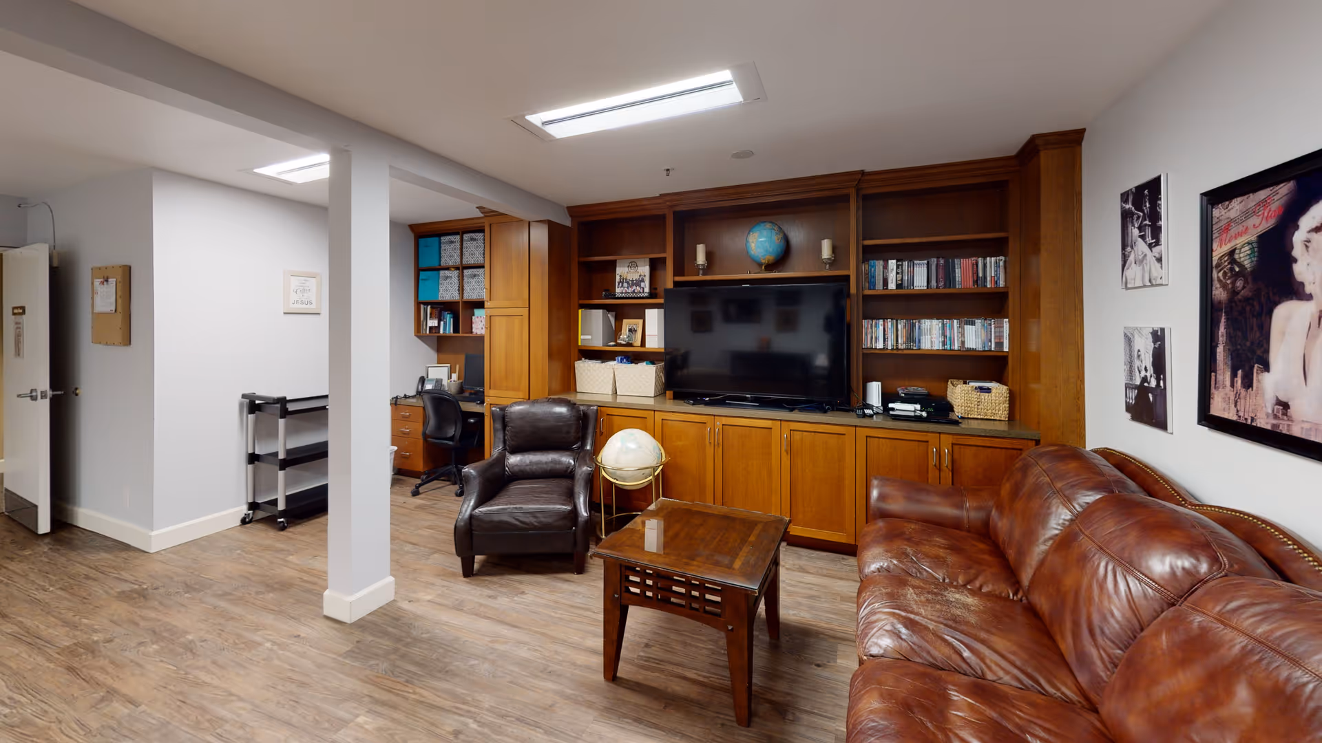 A cozy living room area with a brown leather sofa, a dark brown leather armchair, and a wooden coffee table. Behind the seating is a large wooden built-in shelving unit with a flat-screen TV, books, DVDs, a globe, and decorative items. The room has wood flooring and white walls with framed artwork. To the left, there is a small office nook with a desk and chair.
