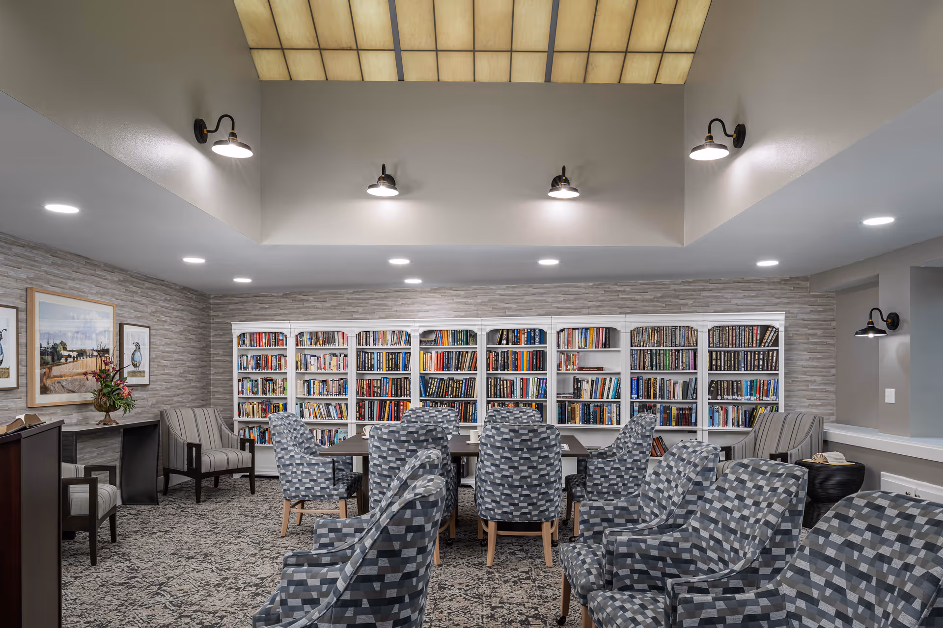A well-lit library room with multiple bookshelves filled with books along the back wall. Several patterned armchairs are arranged around a central table, with additional seating along the sides. The room features a high ceiling with recessed lighting and wall-mounted lamps, and framed artwork is displayed on the left wall.