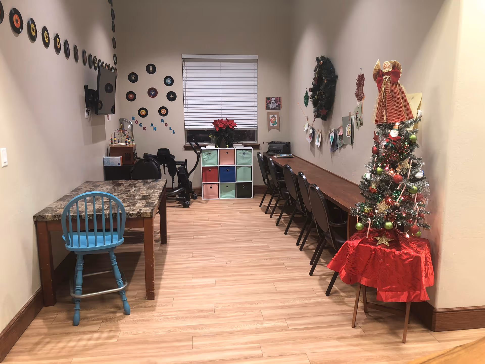 A narrow room with light wood flooring and beige walls decorated with small vinyl records and the words 'Rock and Roll' on one wall. There is a small table with a blue chair on the left side and a long wooden table with several black chairs on the right side. A small decorated Christmas tree with an angel on top is placed on a red cloth-covered stand near the right wall. At the far end, there is a window with closed blinds, a colorful cubby storage unit, and some wall decorations including a wreath and pictures.