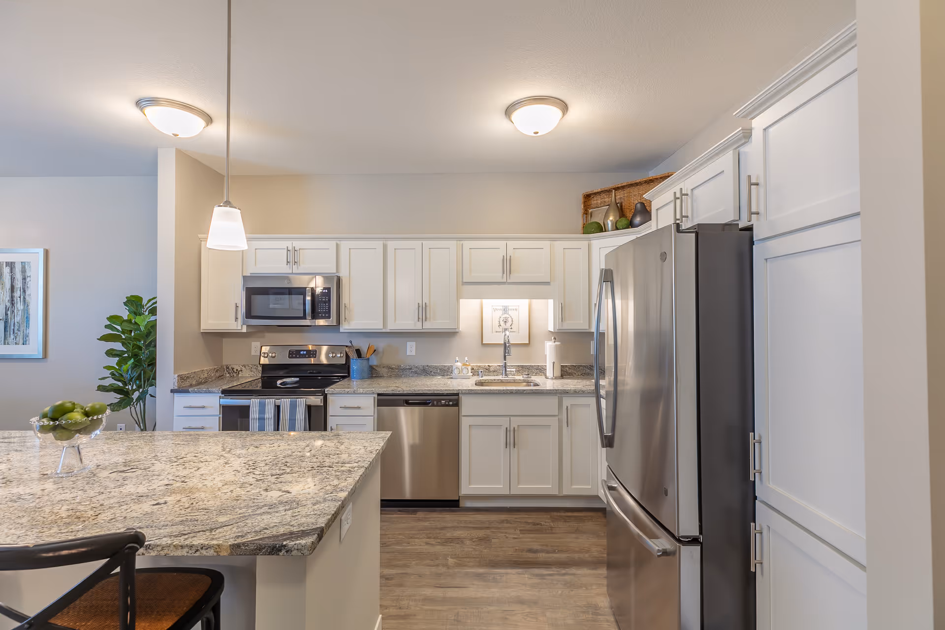 Modern kitchen with white cabinets, granite countertops, stainless steel appliances including a refrigerator, dishwasher, stove, and microwave. There is a kitchen island with a bowl of green apples and a bar stool. The kitchen is well-lit with ceiling lights and a hanging pendant light.