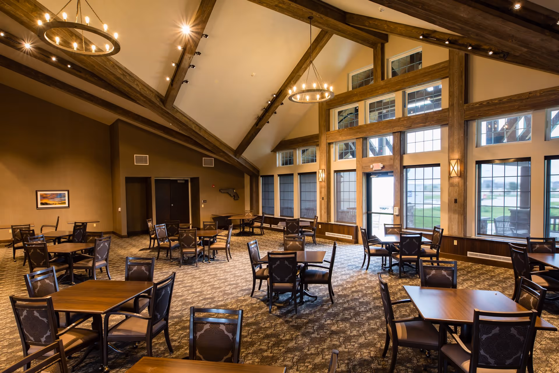 Spacious dining room with multiple wooden tables and chairs arranged on a patterned carpet. The room features high vaulted ceilings with exposed wooden beams and large windows allowing natural light to fill the space. Two circular chandeliers hang from the ceiling, and there is a door leading outside.