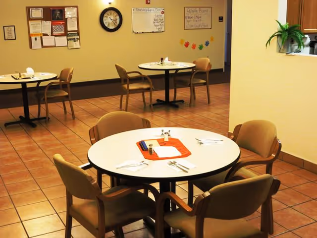 A dining area with round tables and chairs arranged around them. Each table has place settings with napkins, utensils, and salt and pepper shakers. The room has tiled flooring, a bulletin board, a clock on the wall, and some decorative handprints on the far wall. There is a small plant on a counter to the right.