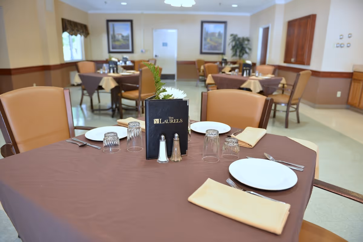 A dining room with tables covered in brown tablecloths and set with white plates, utensils, glasses, and beige napkins. One table in the foreground has a menu labeled 'The Laurels' along with salt and pepper shakers and a small flower arrangement. The room has beige walls with wood trim, framed artwork, and several chairs around each table.
