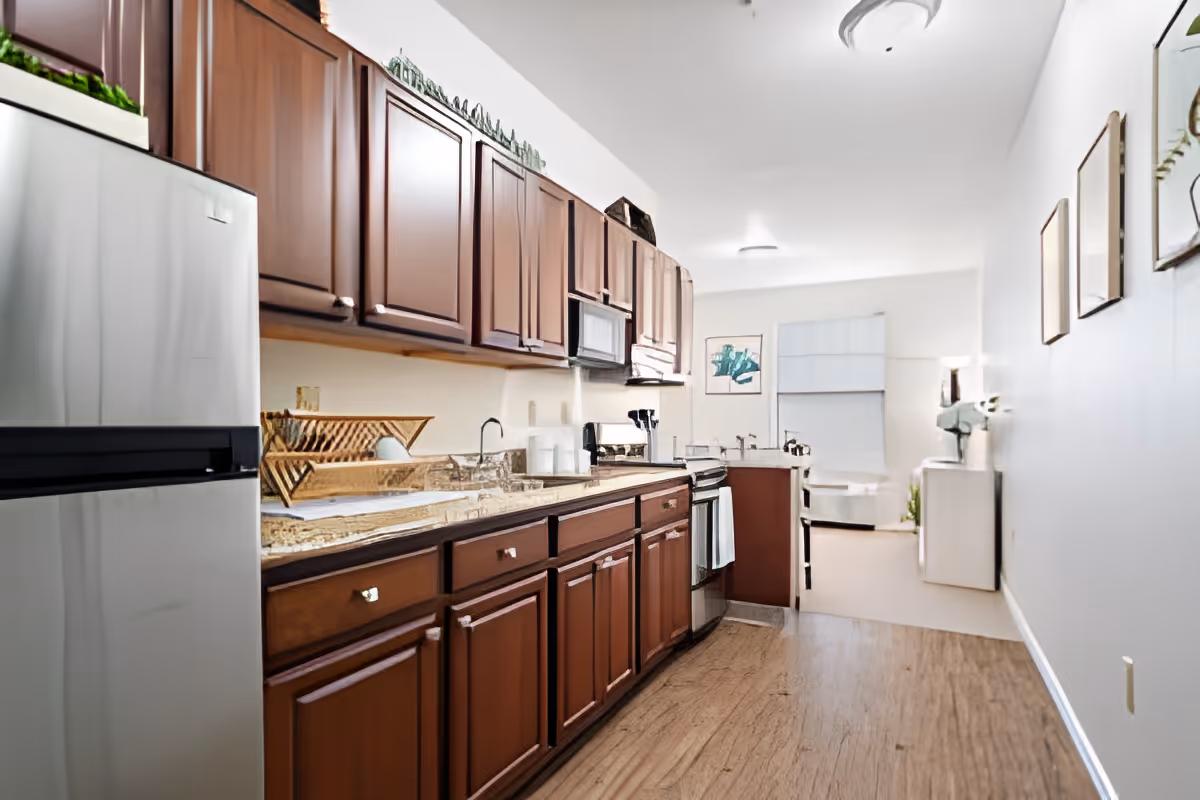 A modern kitchen with wooden cabinets, a stainless steel refrigerator, a microwave, an oven, and a granite countertop. The kitchen opens into a bright living area with a window, artwork on the walls, and a small table with a lamp.