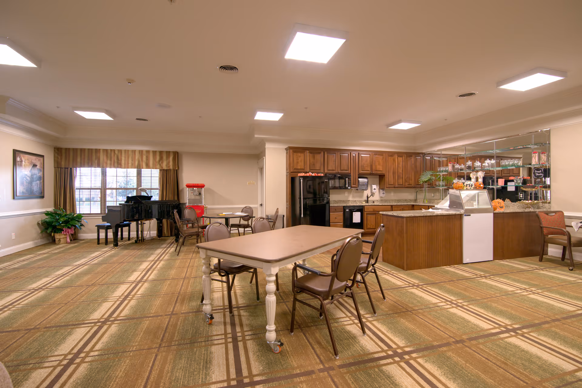 A spacious room with a kitchen area featuring wooden cabinets, a black refrigerator, and a countertop with various items. In the foreground, there are tables and chairs arranged for seating. To the left, there is a black grand piano near a window with striped curtains, and a popcorn machine next to it. The room has a patterned carpet and bright ceiling lights.