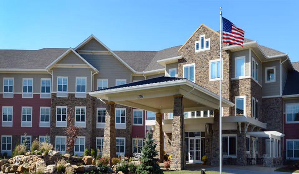 Exterior view of Cedarhurst of Columbia, a multi-story senior living facility with a stone and siding facade, a covered entrance supported by stone pillars, landscaped garden with rocks and plants, and an American flag on a flagpole in front.