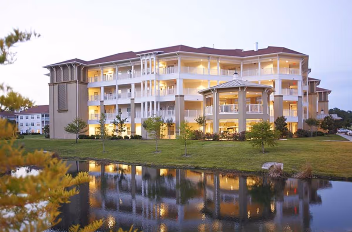 Exterior view of a multi-story senior living facility named The Village at Gainesville, with balconies and warm interior lights reflecting on a pond in the foreground, surrounded by green grass and small trees.
