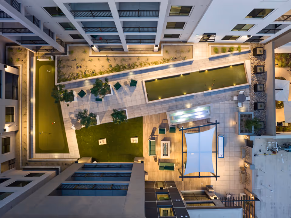Aerial view of an outdoor courtyard area in a senior living facility featuring two bocce ball courts, seating areas with chairs and tables, a shaded canopy over a seating area, greenery, and surrounding building balconies and windows.