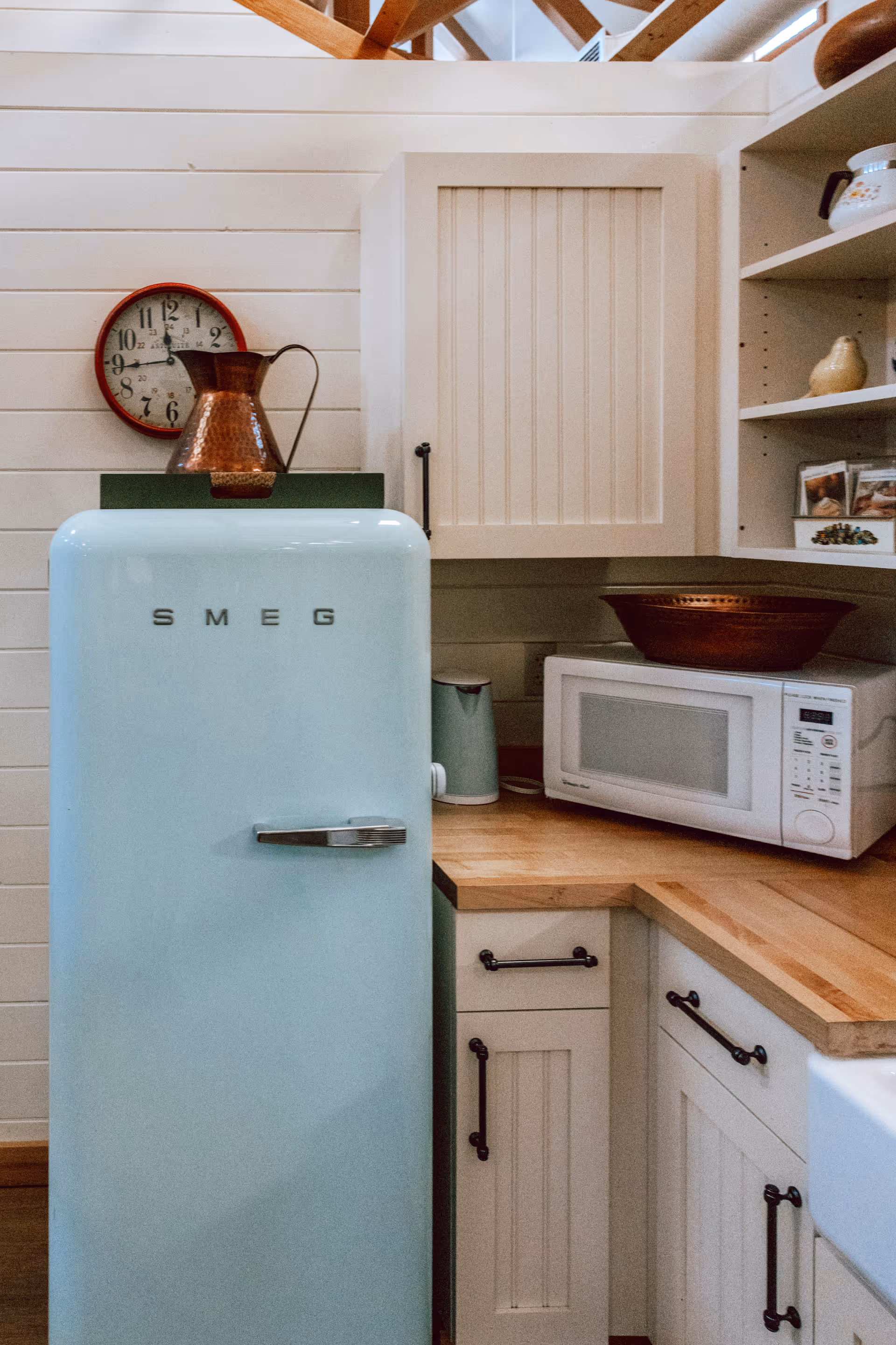 A cozy kitchen corner featuring a light blue SMEG refrigerator, a wooden countertop with a white microwave, a copper pitcher, a clock on the wall, and cream-colored cabinets with black handles.