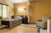 A simple nursing facility bedroom with a single bed, nightstand, armchair, window, and a privacy curtain.