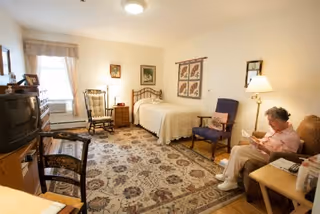 A bright senior living bedroom with a twin bed, armchairs, a TV on a dresser, and a seated resident reading at a side table.