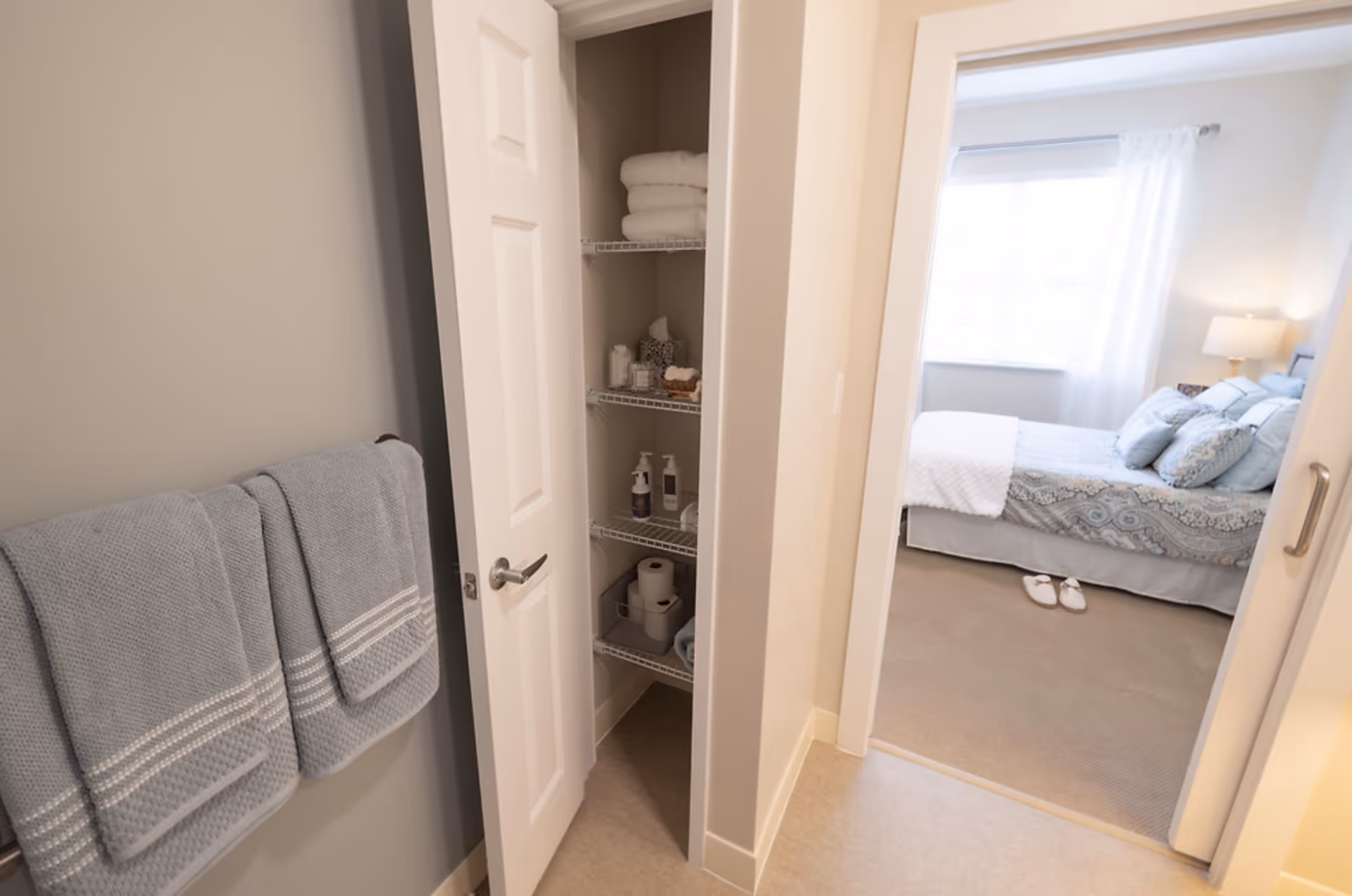 View from a bathroom into a bedroom showing a towel rack, open linen closet with toiletries and towels, and a bed in the adjacent room.