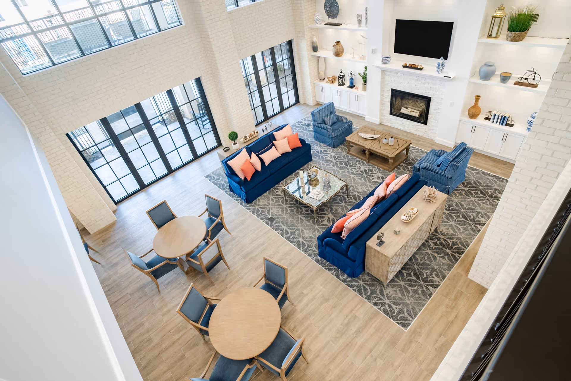 A bright and spacious living room area with large windows and glass doors letting in natural light. The room features two blue sofas with coral and pink cushions, two blue armchairs, a wooden coffee table, and a glass-top side table on a patterned rug. There are built-in white shelves with decorative items and a fireplace with a mounted TV above it. In the foreground, there are two round wooden tables each surrounded by four blue cushioned chairs.