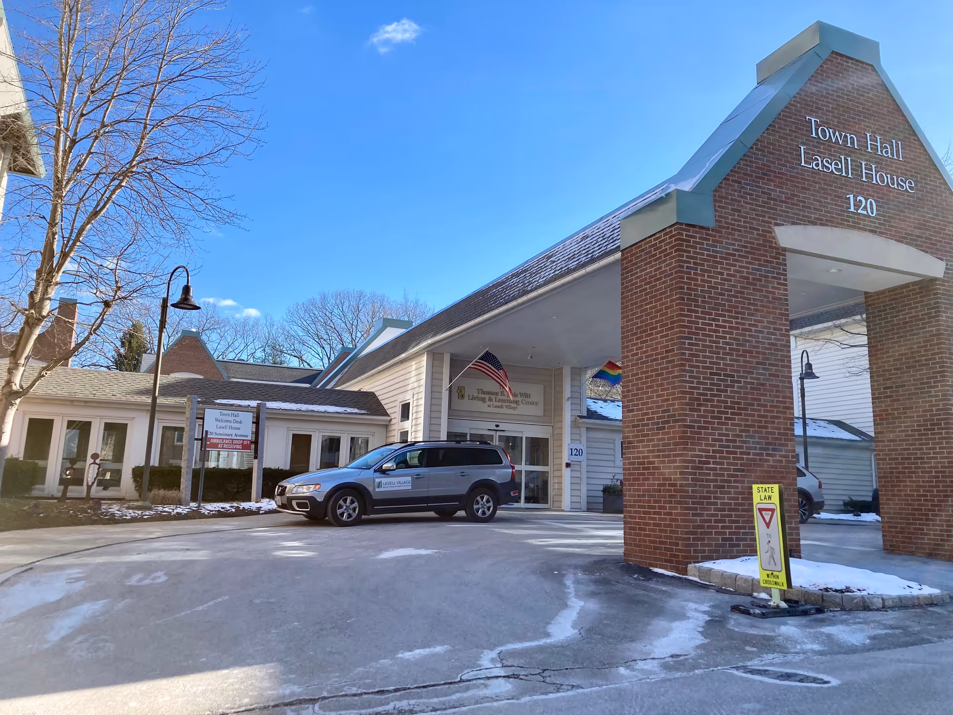 Exterior view of the entrance to Town Hall Lasell House at Lasell Village, showing a brick archway with the building name and number 120. A silver vehicle with a Lasell Village logo is parked near the entrance. There are leafless trees, a street lamp, and a sign indicating ambulance drop off at receiving. The sky is clear and blue.