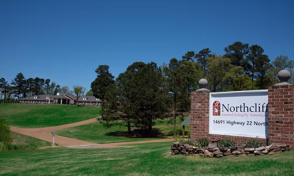 A wide outdoor view of the Northcliff assisted living facility with a large green lawn, a winding driveway, tall trees, and a brick sign displaying the facility's name and address under a clear blue sky.