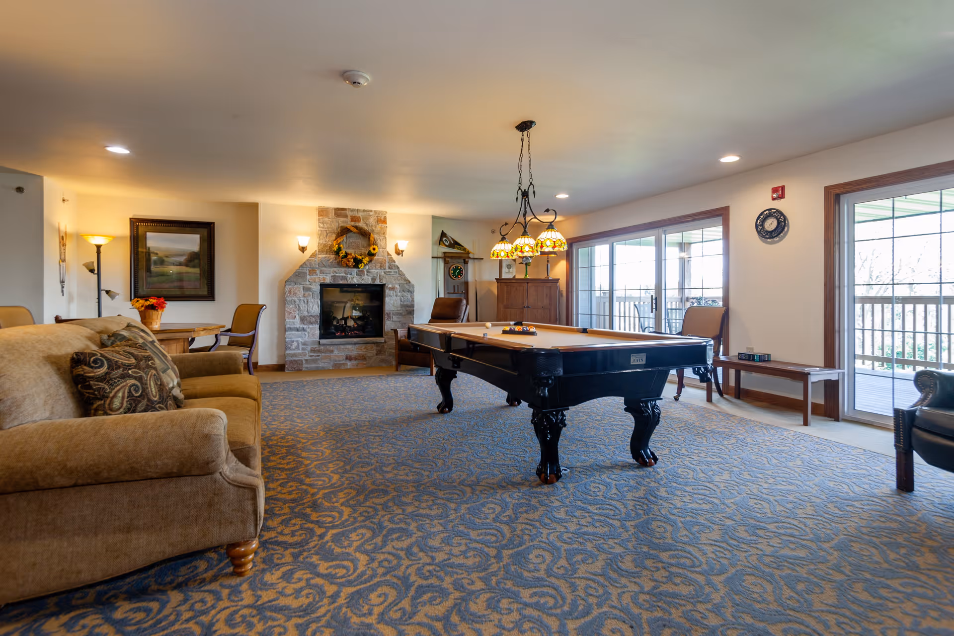 Spacious common lounge with a pool table, sofas, a stone fireplace, and large windows opening to a balcony.