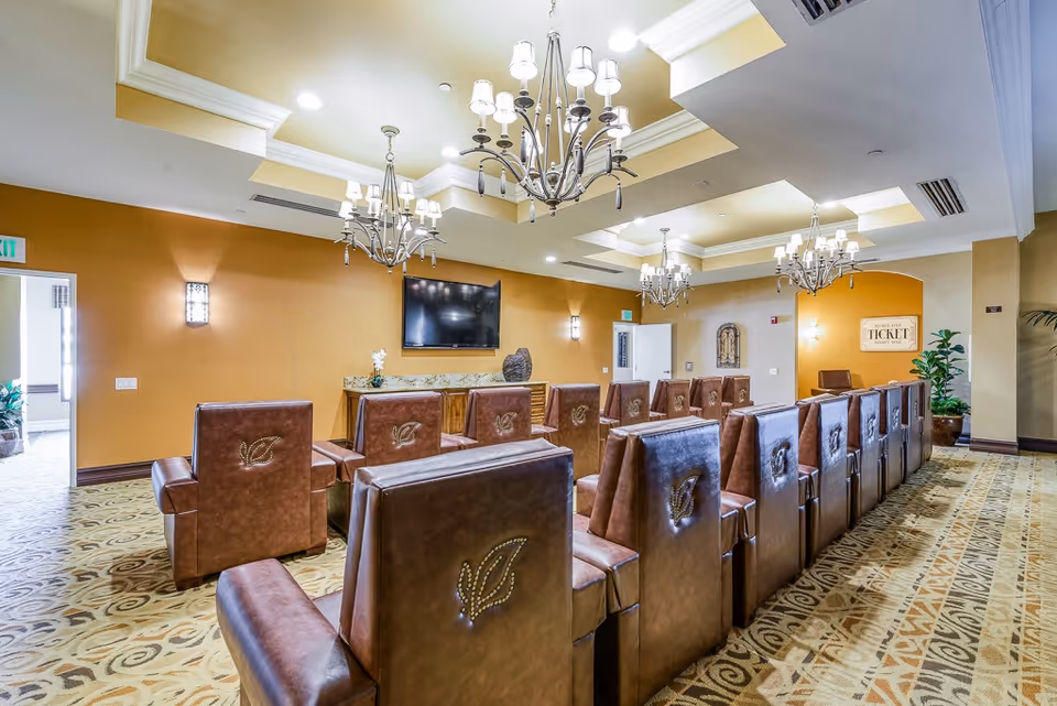 A large room with multiple rows of brown leather chairs facing a wall-mounted flat screen TV. The room has warm yellow walls, patterned carpet, and elegant chandeliers hanging from a tray ceiling. There are decorative plants and a sign on the far wall that reads 'ADMIT ONE TICKET ADMIT YOU'.