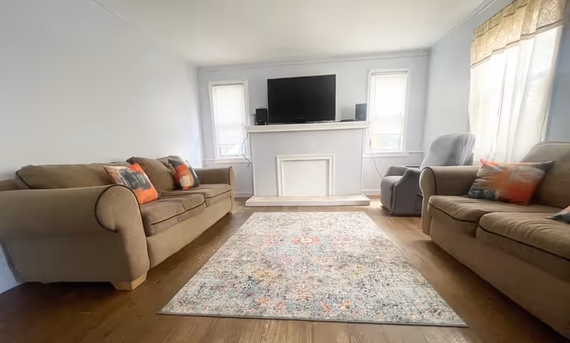 A living room with two brown sofas facing each other, each with colorful throw pillows. A patterned area rug is centered on the wooden floor between the sofas. At the far end of the room is a white fireplace with a flat-screen TV mounted above it, flanked by two windows with blinds. A gray armchair is positioned near the right sofa, next to a window with sheer curtains.