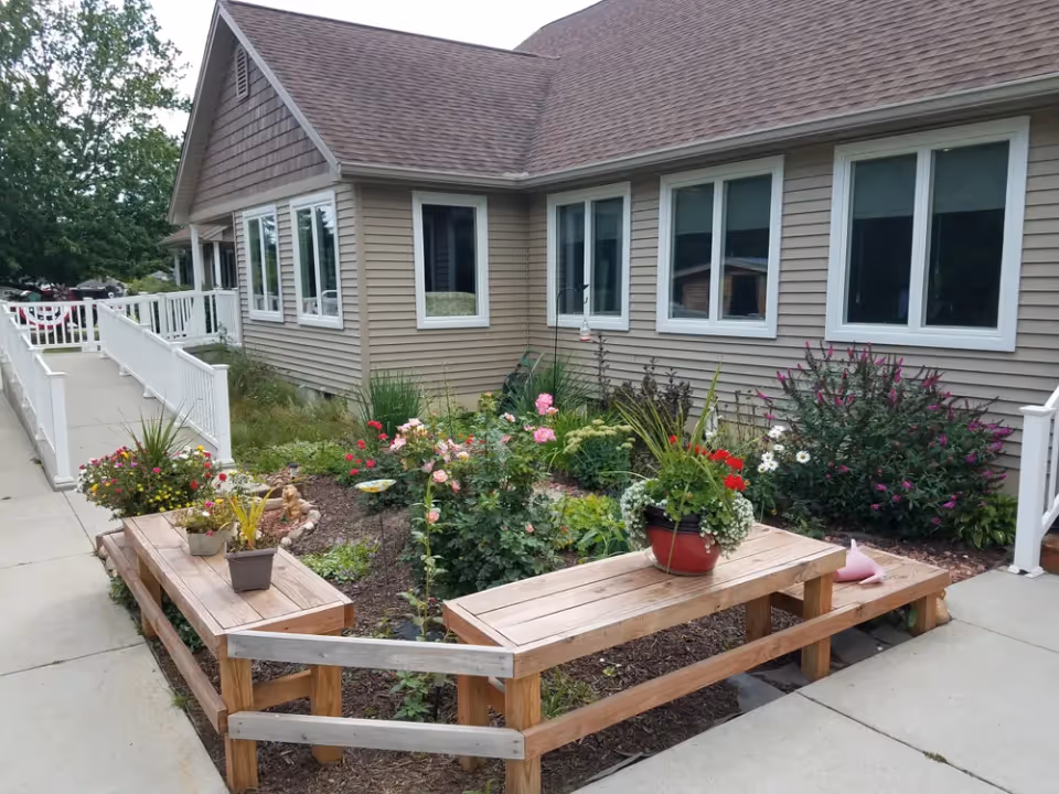 Outdoor garden area at Serenity Senior Living with a variety of flowering plants and shrubs surrounded by wooden benches. The garden is adjacent to a beige building with multiple windows and a ramp leading to the entrance.