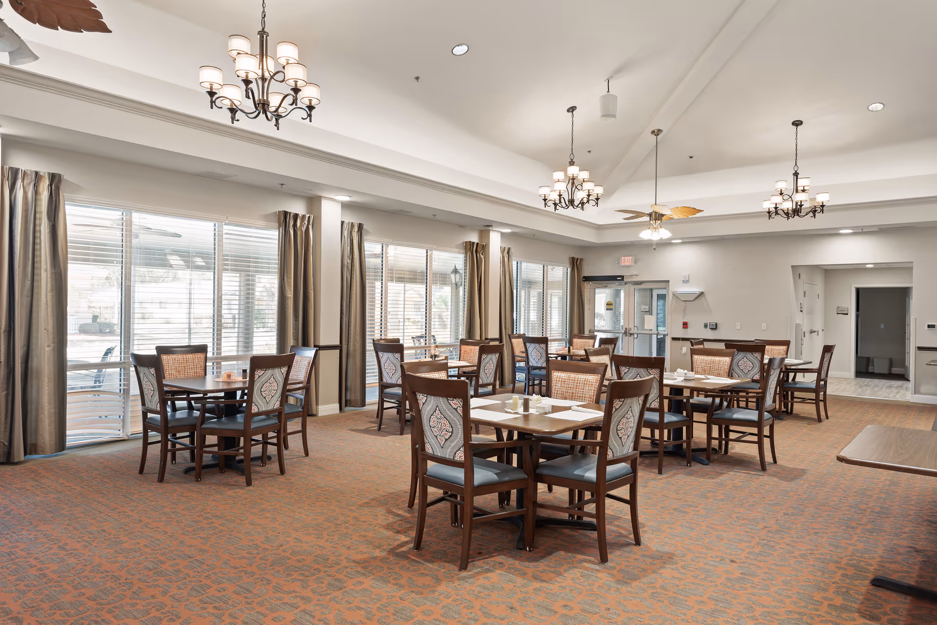 Spacious dining room with multiple wooden tables and chairs, large windows with curtains, and chandeliers overhead.