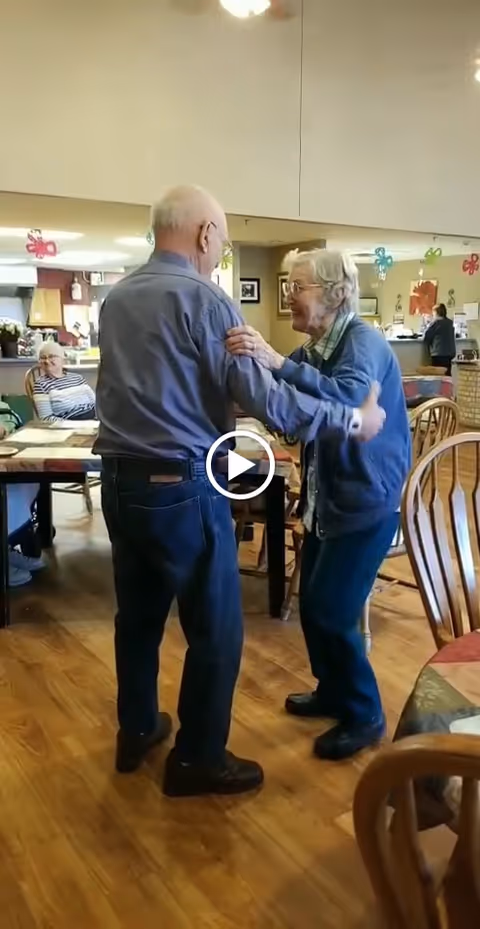 Two elderly men dancing together in a communal dining area with wooden floors and tables. Other seniors are seated at tables in the background, and colorful decorations hang from the ceiling.