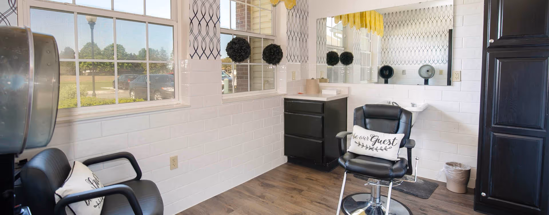 Interior view of a salon area with two black salon chairs, one with a pillow that says 'be our Guest'. There is a large mirror on the wall, a small sink, a black cabinet, and windows showing an outdoor parking lot and greenery.