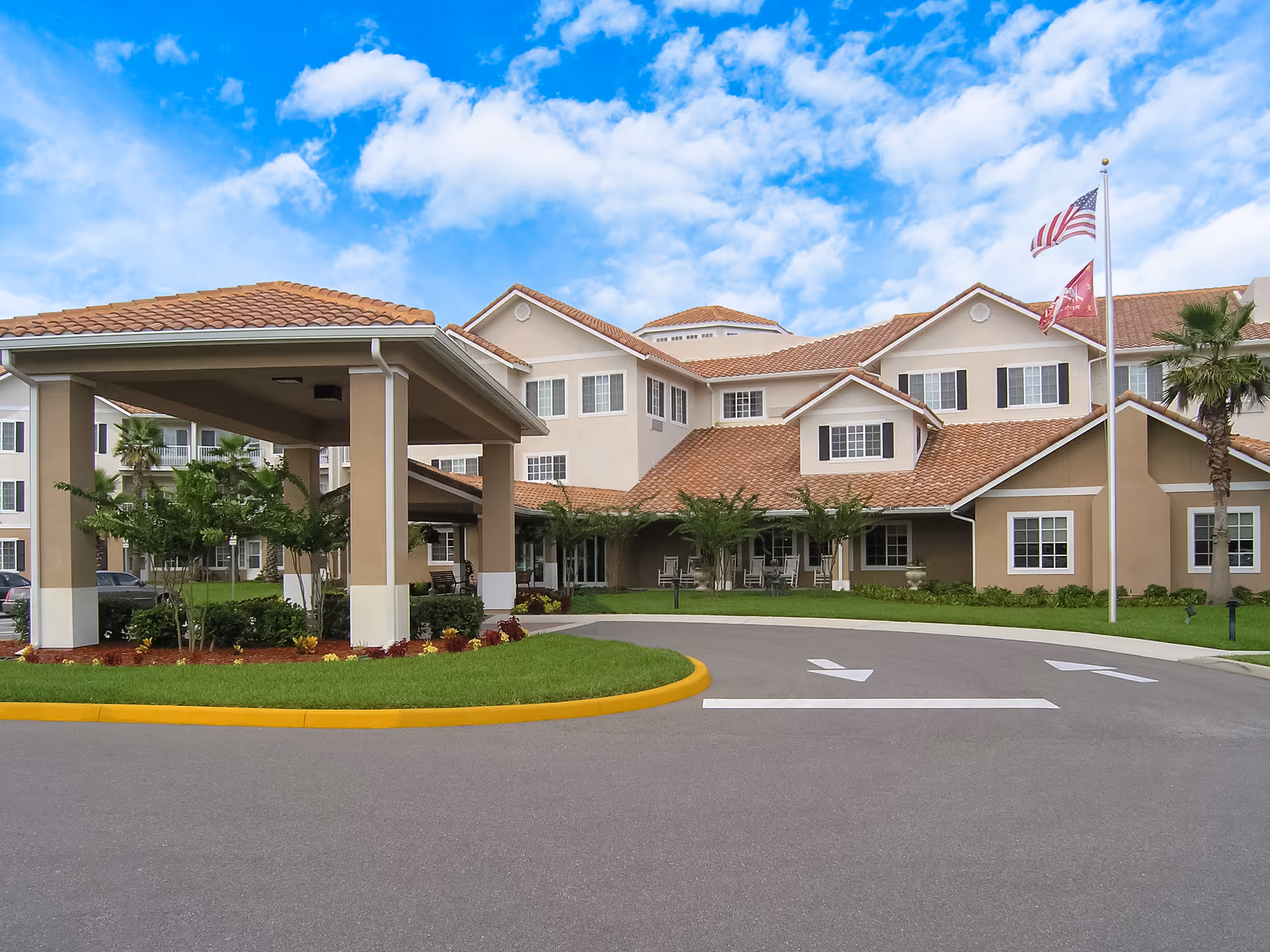 Exterior view of a large senior living facility building with beige walls and a red-tiled roof under a partly cloudy blue sky. There is a covered driveway entrance with columns, landscaped greenery, and two flagpoles flying the American flag and another flag.