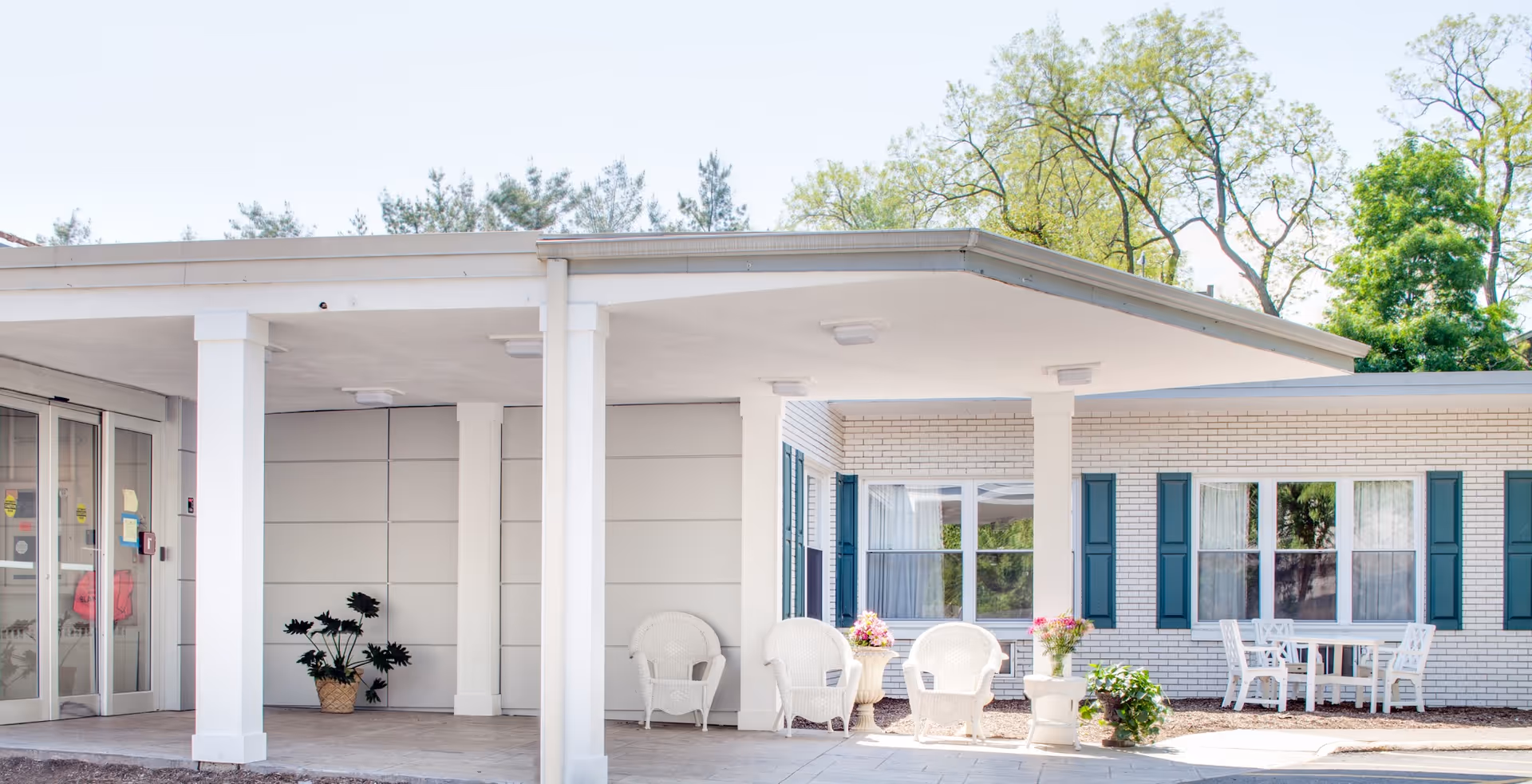 Covered front entrance of a senior living building with white columns, wicker chairs, potted plants and windows with teal shutters.