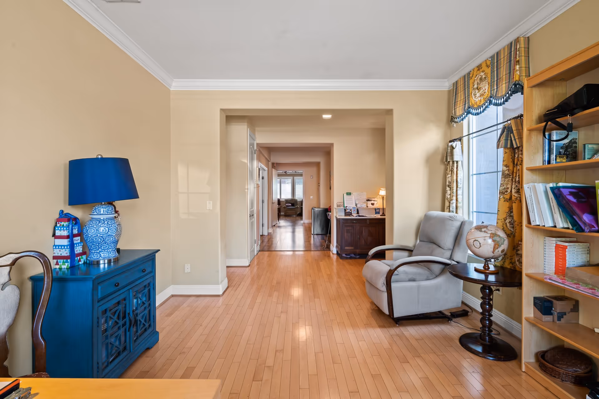 Interior view of a senior living facility room with light wood flooring, beige walls, and white crown molding. On the left, there is a blue cabinet with a blue and white ceramic lamp and a decorative gift bag. On the right, a gray armchair sits next to a round wooden side table with a globe on it, near a window with patterned yellow curtains. A wooden bookshelf filled with books and items is also visible. The hallway extends into another room with more seating visible in the distance.