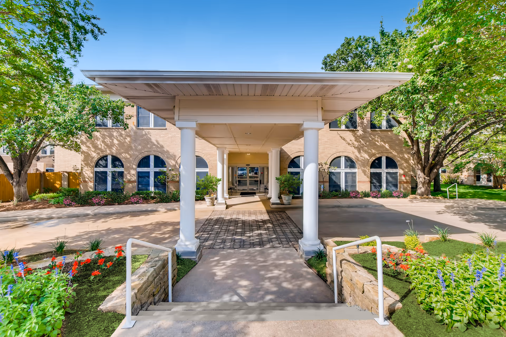 Entrance to Asher Point Senior Living of Austin featuring a covered walkway supported by white columns, surrounded by landscaped gardens with colorful flowers and trees, leading to glass doors of a beige brick building with arched windows.