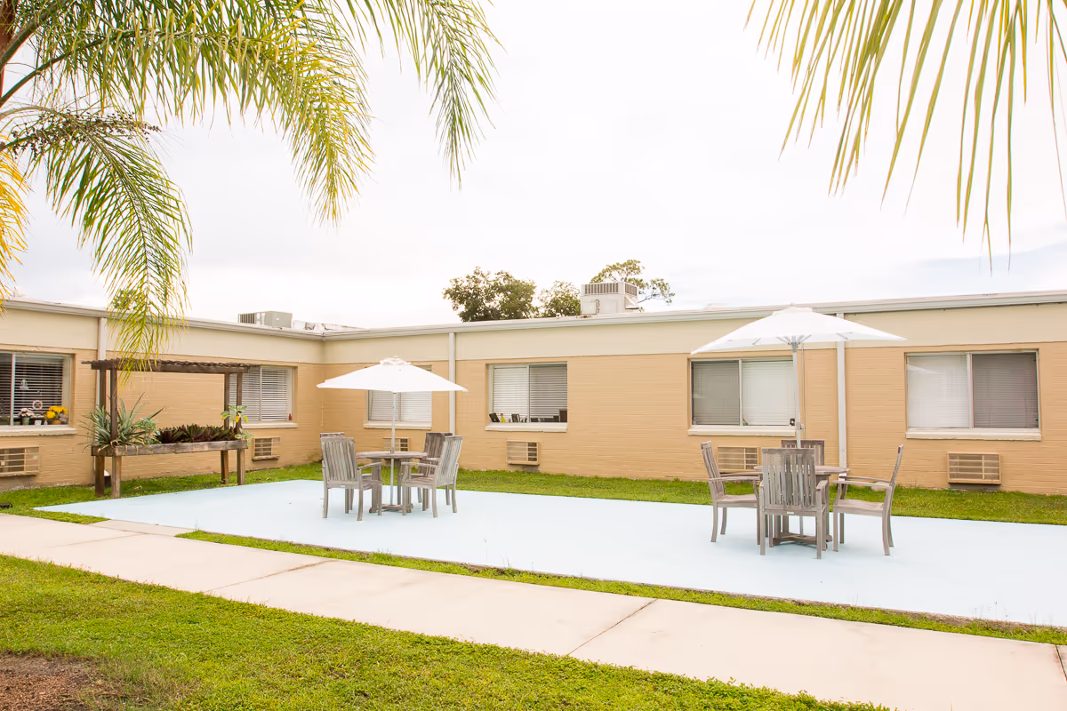 Outdoor courtyard featuring patio tables with umbrellas, grass and palm fronds in front of a single-story beige care facility.