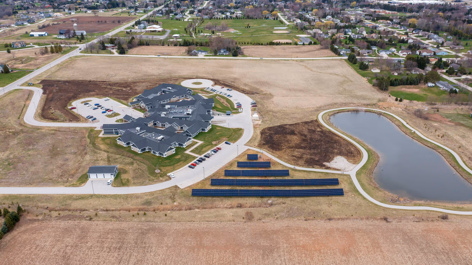 Aerial view of a senior living campus showing a cluster of connected buildings, parking lots, rows of solar panels, and a nearby pond set in open fields.