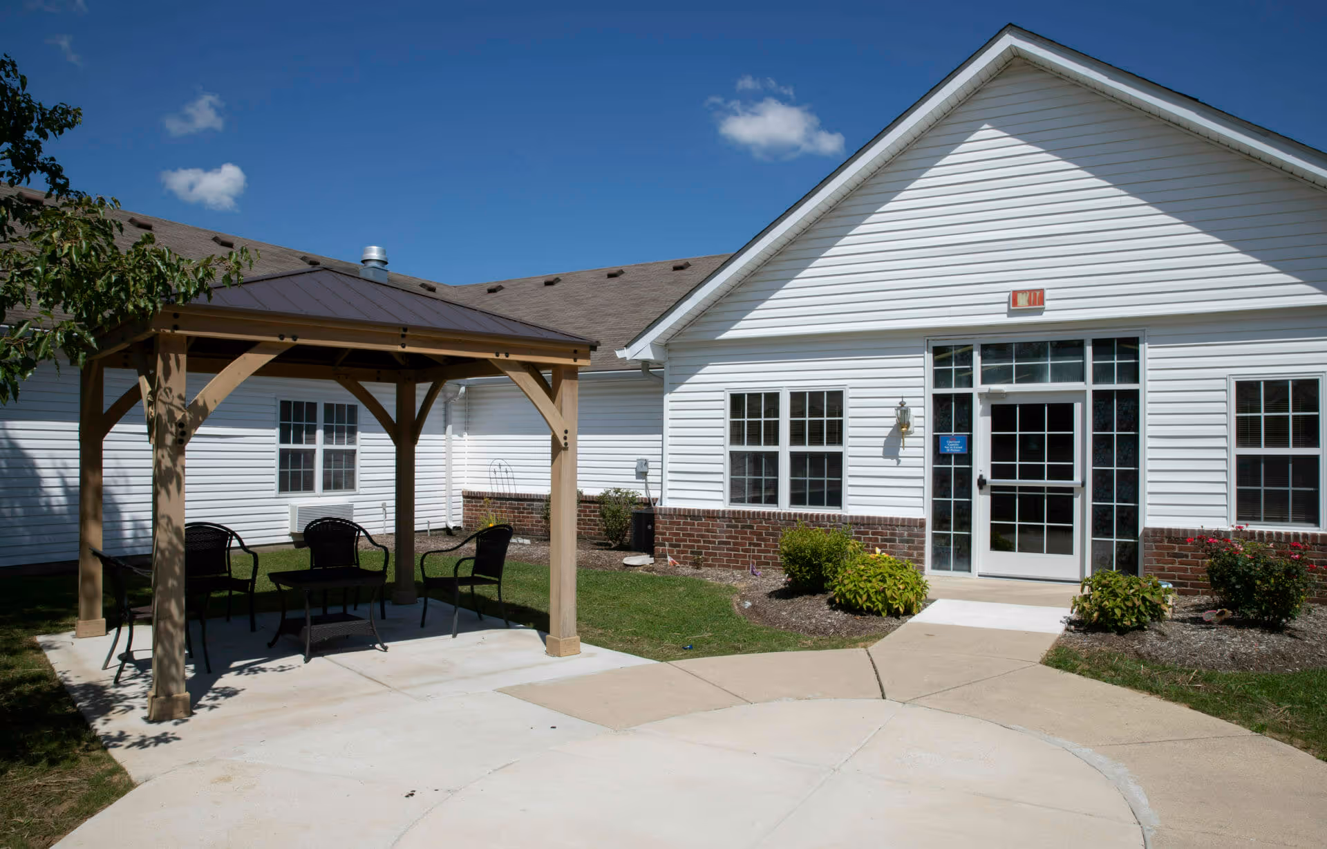 Outdoor patio area at Brownsburg Meadows featuring a wooden gazebo with four black chairs and a small table underneath. The building exterior is white with brick accents and multiple windows. A concrete walkway leads to a glass door entrance under a clear blue sky.