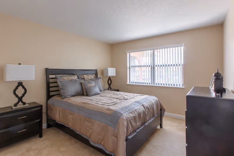 Bright bedroom with a made bed, two nightstands with lamps, a dresser, and a window with vertical blinds.