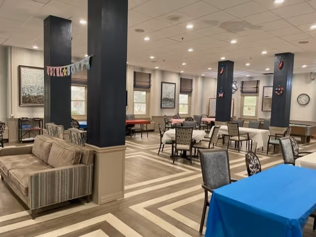A spacious common area with multiple tables and chairs arranged for dining or activities. The room features patterned flooring, several black pillars decorated with small wreaths and banners, and a beige couch near one of the pillars. The walls have framed artwork and windows with blinds, and the ceiling has recessed lighting.