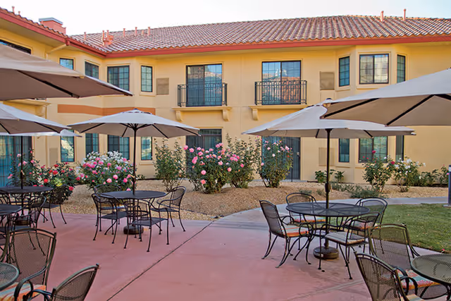 Courtyard patio with metal tables, chairs and umbrellas in front of a two-story yellow stucco building with rose bushes.