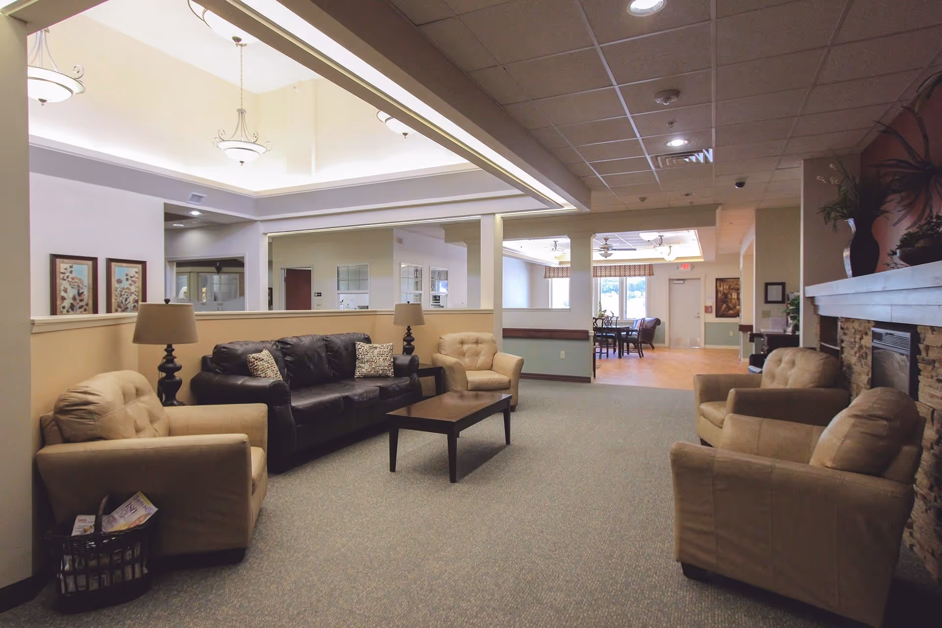 A cozy living room area in a senior living facility with beige armchairs and a dark brown leather sofa arranged around a wooden coffee table. The room features soft lighting from ceiling fixtures, a stone fireplace on the right, and a dining area visible in the background through an open partition.