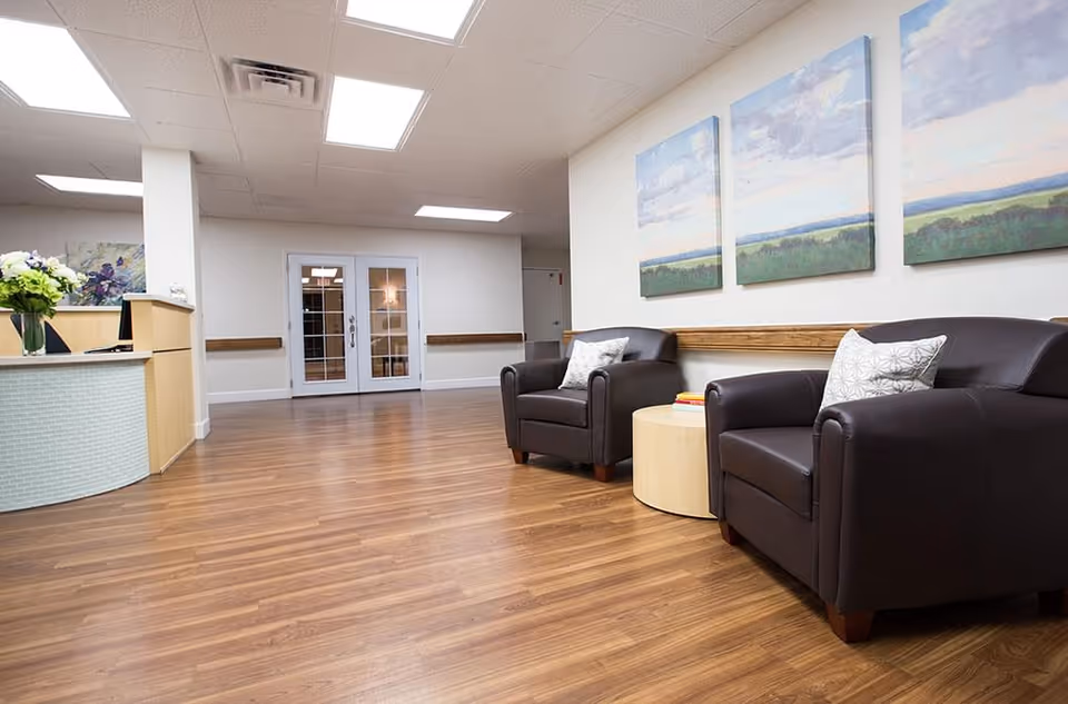 A bright and clean waiting area with two dark brown leather armchairs, a round wooden side table with books, and three landscape paintings on the wall. The floor is wood laminate, and there is a reception desk with a flower vase on the left side. Double glass doors are visible in the background.