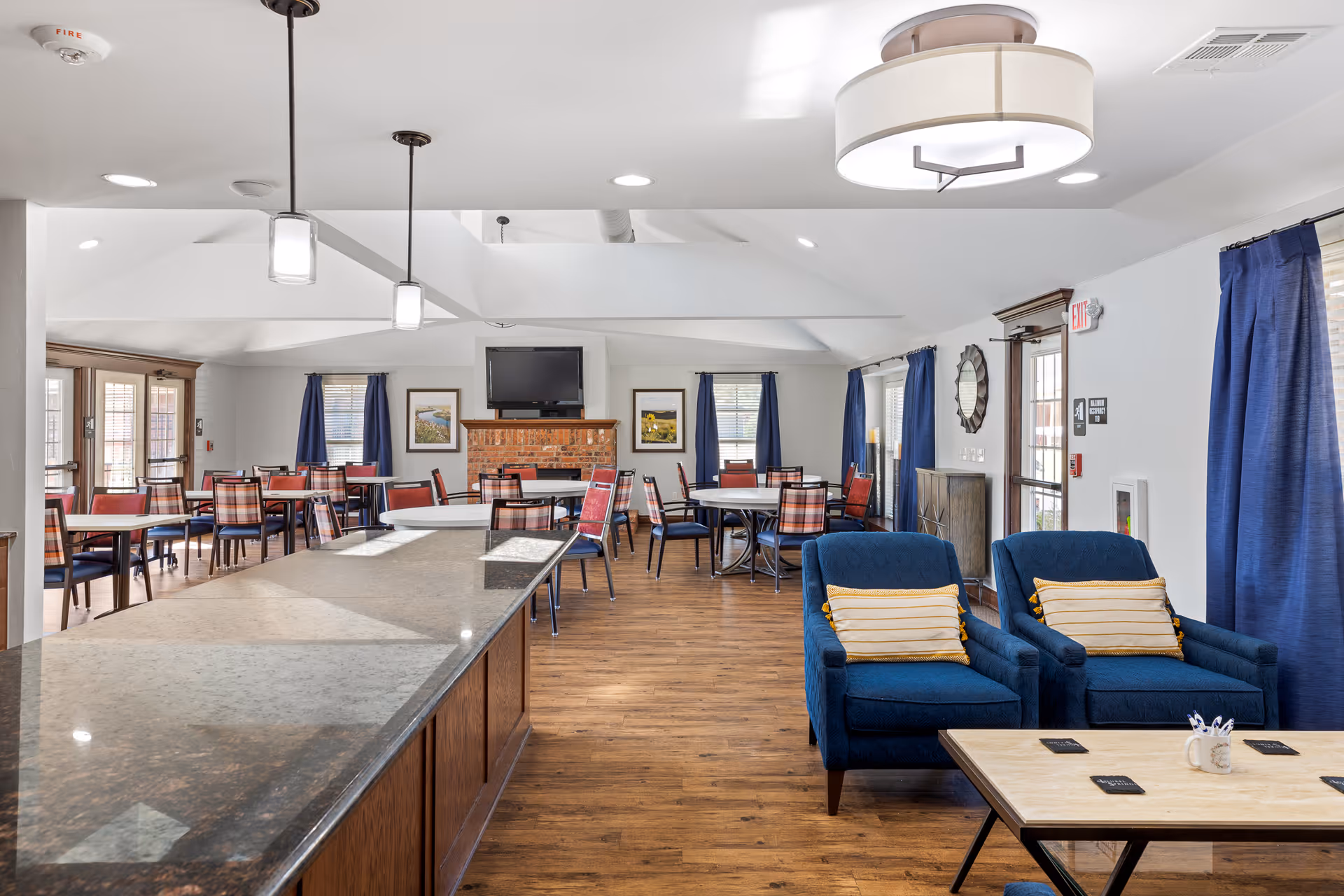 Bright communal dining and lounge area with multiple dining tables and chairs, a TV above a brick fireplace, a long kitchen island, and two blue armchairs.